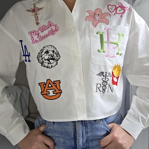 May include: White button-down shirt with various embroidered patches. Patches include a cross, a dog, the text "My Black Is Beautiful", a floral design, a baseball team logo, initials, and a medical symbol with fries.