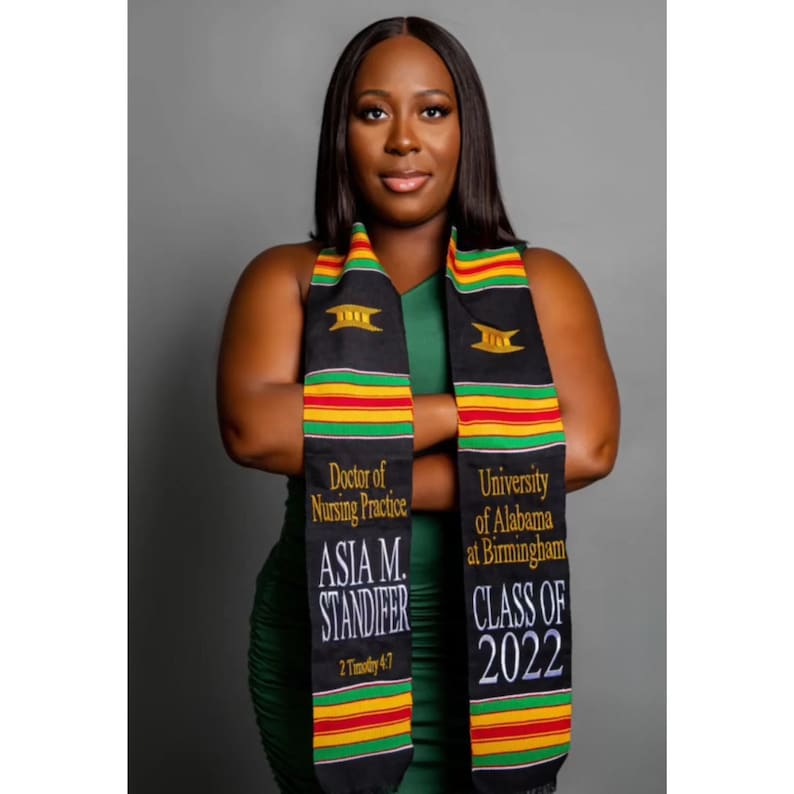 May include: A woman wearing a green dress and a graduation stole. The black stole has gold, red, and green stripes with the text "Doctor of Nursing Practice," "ASIA M. STANDIFER," and "CLASS OF 2022." The background is a solid gray.