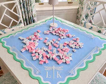 May include: A light blue square mahjong mat with scalloped green and white edges, featuring a white dragonfly graphic and the letters L K. The mat includes clear acrylic corner guards and is covered in scattered pink and white mahjong tiles.