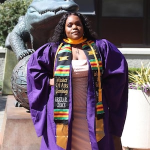 May include: A person wearing a purple graduation gown and a gold and black sash that reads "Master of Arts Gerontology Ursula R. Choice." The person is standing in front of a statue of an alligator.