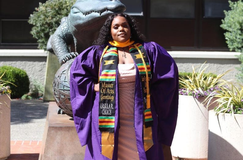 May include: A person wearing a purple graduation gown and a gold and black sash that reads "Master of Arts Gerontology Ursula R. Choice." The person is standing in front of a statue of an alligator.