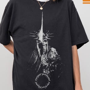 以下が含まれることがあります： 黒いTシャツ。王冠、剣、指輪を持った骸骨の白いグラフィックが描かれています。「Comfort Colors」の文字が右上。Tシャツは黒で、左上に「COLOR: BLACK」の文字があります。