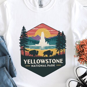 Könnte beinhalten: Weißes T-Shirt mit einem Vintage-Grafikdruck des Yellowstone Nationalparks. Die Grafik zeigt einen ausbrechenden Geysir, einen Sonnenuntergang und zwei Bisons. Der Text "Yellowstone National Park" ist unter der Grafik gedruckt.