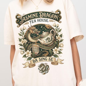 Pode incluir: Camiseta cor marfim com um gráfico detalhado. O design inclui um dragão verde, uma chaleira e o texto "Jasmine Dragon Tea House" e "Ba Sing Se". O gráfico é rodeado por elementos florais.