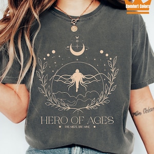 May include: Pepper-colored t-shirt featuring a celestial design with a figure, crescent moon, and stars. The text "HERO OF AGES" and "THE MISTS ARE MINE" are printed below the graphic. The shirt is a Comfort Colors brand.