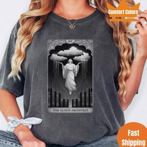 May include: A grey t-shirt featuring a black and white graphic of a tarot card. The card is labelled "The Cloud Architect" and depicts a figure with outstretched arms standing in front of a cloud containing a city. The card number is XII.
