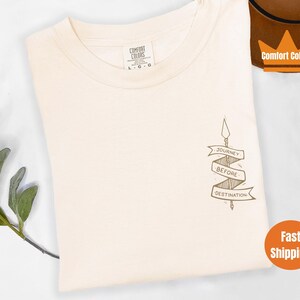 Puede incluir: Una camiseta de color crema con un gr&aacute;fico marr&oacute;n de una flecha con una pancarta que dice "Journey Before Destination".