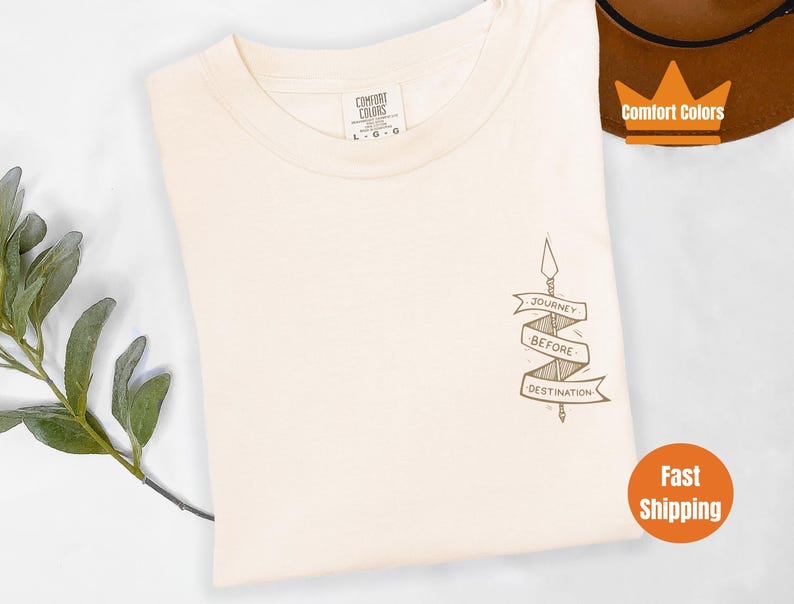 Puede incluir: Una camiseta de color crema con un gr&aacute;fico marr&oacute;n de una flecha con una pancarta que dice "Journey Before Destination".