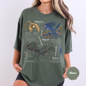 May include: A moss green crewneck t-shirt featuring a detailed graphic print of three dragons labeled Andarna, Sgaeyl, and Tairn. The design includes swords, feathers, and descriptive text in a fantasy style, with a circular color swatch labeled Moss.