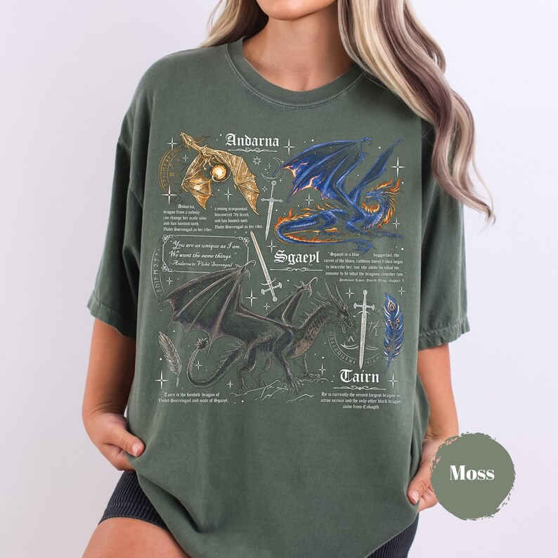 May include: A moss green crewneck t-shirt featuring a detailed graphic print of three dragons labeled Andarna, Sgaeyl, and Tairn. The design includes swords, feathers, and descriptive text in a fantasy style, with a circular color swatch labeled Moss.