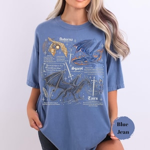 May include: A blue short-sleeve t-shirt featuring a detailed graphic print of three dragons labeled Andarna, Sgaeyl, and Tairn. The design includes intricate illustrations of swords, feathers, and text descriptions in a vintage style font.