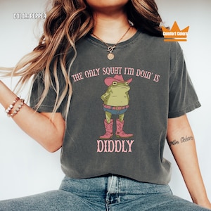May include: Pepper-colored t-shirt featuring a cartoon frog in a cowboy hat and boots. The text reads "THE ONLY SQUAT I'M DOIN' IS DIDDLY" in pink. The shirt is a Comfort Colors brand.