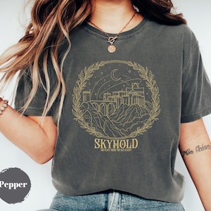 May include: Dark grey t-shirt with a gold illustration of a city, mountains, and a crescent moon inside a laurel wreath. The word "SKYHOLD" and the phrase "THE PLACE WHERE THE SKY IS KEPT" are printed below the illustration. The shirt is labeled "Pepper".