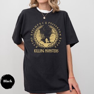 May include: Black t-shirt with a gold graphic featuring a forest scene, a crescent moon, and the text "KILLING MONSTERS". The design is encircled by a wreath and runes. The shirt is worn by a person.