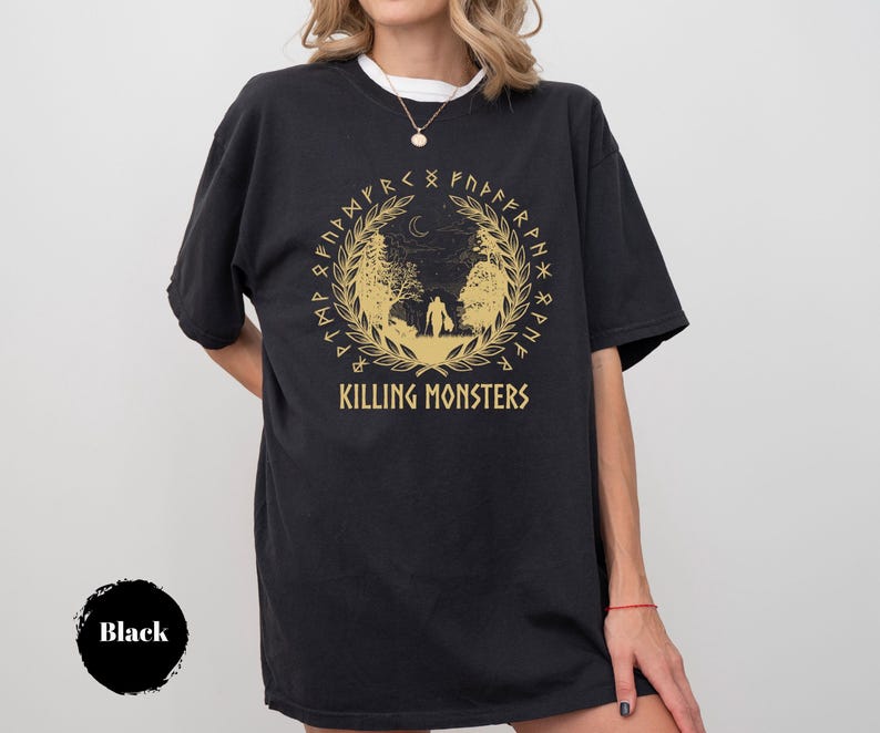 May include: Black t-shirt with a gold graphic featuring a forest scene, a crescent moon, and the text "KILLING MONSTERS". The design is encircled by a wreath and runes. The shirt is worn by a person.