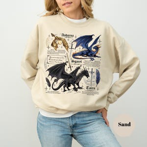 May include: A sand-colored crewneck sweatshirt featuring a graphic print of three dragons labeled Andarna, Sgaeyl, and Tairn. The design includes detailed illustrations of the dragons, swords, feathers, and small text blocks surrounding each creature.
