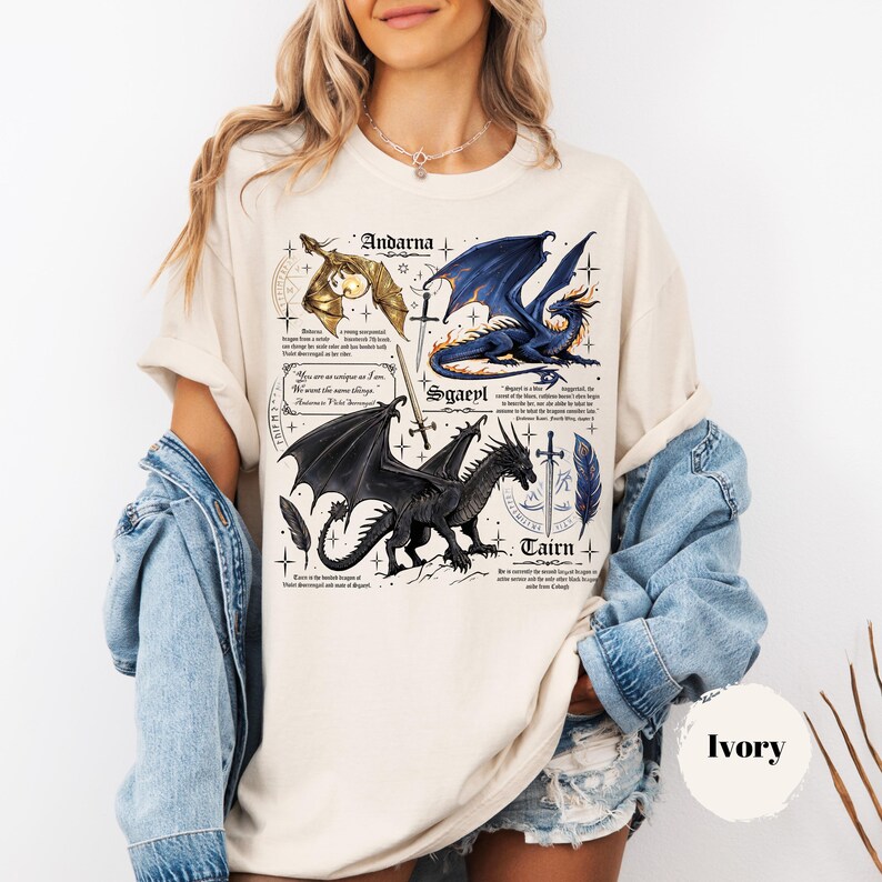 May include: An ivory t-shirt featuring a detailed graphic print of three dragons labeled Andarna, Sgaeyl, and Tairn. The design includes illustrations of swords, feathers, and small text blocks describing each dragon, set against a light background.