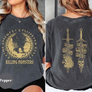 May include: Pepper-colored t-shirt with a gold graphic on the front and back. The front design features a forest scene with the text "KILLING MONSTERS" and runes. The back displays two swords with the text "MONSTER SLAYER" and "MAN SLAYER".