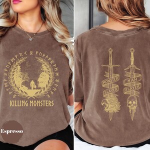 May include: Espresso-colored t-shirt with a gold graphic on the front featuring a forest scene and the text "KILLING MONSTERS." The back displays two swords with the text "MONSTER SLAYER" and "MAN SLAYER."