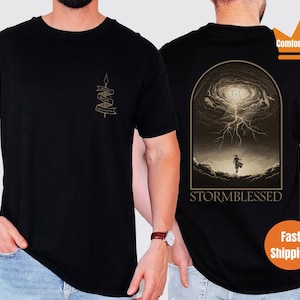 Puede incluir: Camiseta negra con un gr&aacute;fico de una persona de pie en un cielo tormentoso con el texto "STORMBLESSED" debajo. La camiseta tiene un peque&ntilde;o gr&aacute;fico en el pecho izquierdo con el texto "Storm Blessed" en una pancarta.