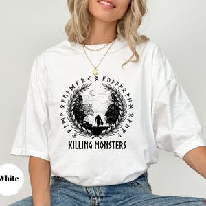 May include: White t-shirt with a black graphic featuring a silhouette of a person in a forest, surrounded by runes and the text "KILLING MONSTERS". The design includes a crescent moon and laurel wreath.
