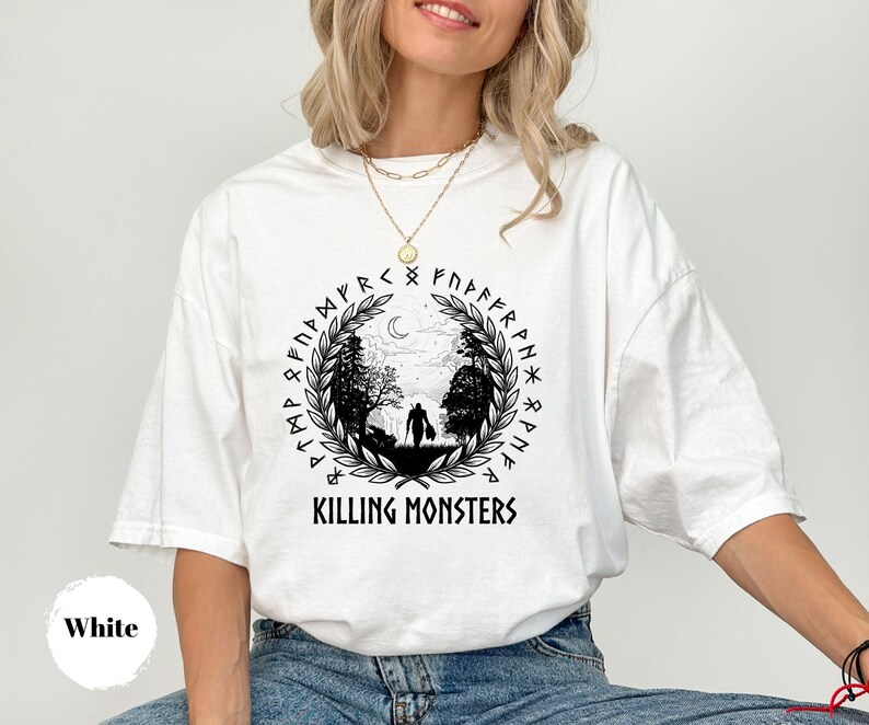 May include: White t-shirt with a black graphic featuring a silhouette of a person in a forest, surrounded by runes and the text "KILLING MONSTERS". The design includes a crescent moon and laurel wreath.