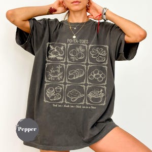 Op de afbeelding: Donkergrijs T-shirt met een raster van aardappelillustraties en de tekst "PO-TA-TOES". Daaronder staat "Boil 'em • Mash 'em • Stick 'em in a Stew". Het T-shirt heeft een losse pasvorm.