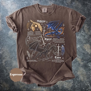 May include: An espresso-colored short-sleeve t-shirt featuring a graphic print of three dragons labeled Andarna, Sgaeyl, and Tairn. The design includes detailed illustrations of dragons, swords, feathers, and small text excerpts from the Fourth Wing book series.