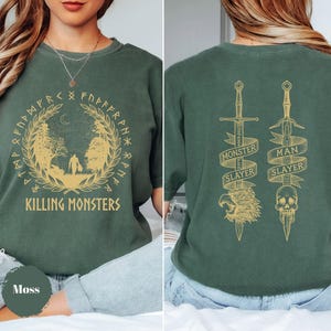 May include: Moss green t-shirt with a gold design featuring the text "KILLING MONSTERS" and a circular illustration. The back displays two swords with the text "MONSTER SLAYER" and "MAN SLAYER".