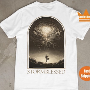 Puede incluir: Camiseta blanca con un estampado gr&aacute;fico de una persona caminando hacia una nube de tormenta con rayos. El texto "STORMBLESSED" est&aacute; impreso debajo del gr&aacute;fico.