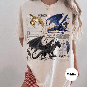 May include: A cream-colored t-shirt featuring a graphic print of three dragons labeled Andarna, Sgaeyl, and Tairn. The design includes illustrations of swords, feathers, and text excerpts. The shirt has short sleeves and a crew neckline.