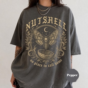 May include: Dark gray t-shirt with a distressed graphic. The design features a moth with a skull on its body, a crescent moon, and the words "NUTSHELL" and "no place to call home" in a vintage font. The shirt is labeled "Pepper".