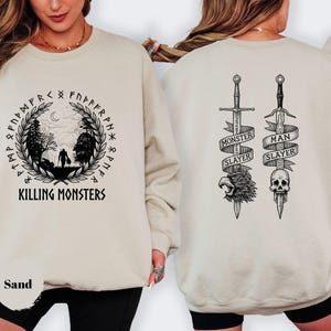 May include: Sand-colored sweatshirt with a black graphic on the front featuring a figure walking through a forest, surrounded by runes and the text "KILLING MONSTERS." The back displays two swords with banners reading "MONSTER SLAYER" and "MAN SLAYER."