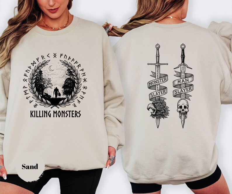 May include: Sand-colored sweatshirt with a black graphic on the front featuring a figure walking through a forest, surrounded by runes and the text "KILLING MONSTERS." The back displays two swords with banners reading "MONSTER SLAYER" and "MAN SLAYER."