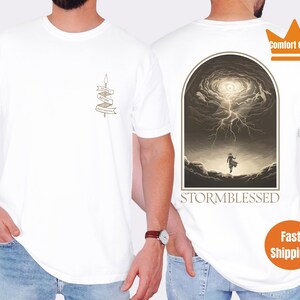 Puede incluir: Camiseta blanca con un gr&aacute;fico de una persona de pie en un cielo tormentoso con el texto "Stormblessed" debajo. La camiseta tambi&eacute;n tiene un peque&ntilde;o gr&aacute;fico de una pancarta con el texto "Stormblessed" en ella.