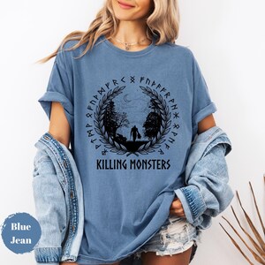 May include: Blue jean colored t-shirt with a circular graphic. The graphic features a silhouette of two figures walking in a forest, surrounded by a wreath and runes. The text "KILLING MONSTERS" is below the graphic.