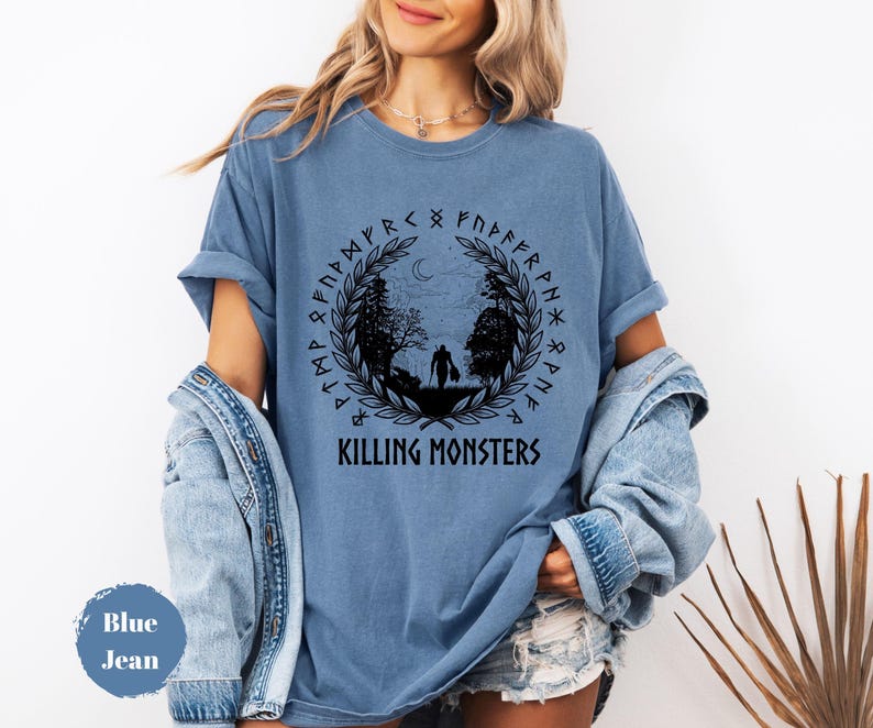 May include: Blue jean colored t-shirt with a circular graphic. The graphic features a silhouette of two figures walking in a forest, surrounded by a wreath and runes. The text "KILLING MONSTERS" is below the graphic.