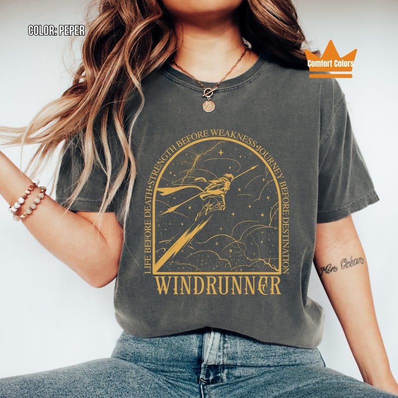 Puede incluir: Camiseta gris oscuro con un gr&aacute;fico dorado de una figura con una lanza, la palabra "WINDRUNNER" y el texto "LIFE BEFORE DEATH - STRENGTH BEFORE WEAKNESS - JOURNEY BEFORE DESTINATION". La camiseta es de la marca Comfort Colors.