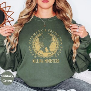 May include: Military green sweatshirt with a gold graphic featuring a forest scene, a crescent moon, and the text "KILLING MONSTERS". The design is encircled by a wreath and runic symbols. The sweatshirt is a solid color.