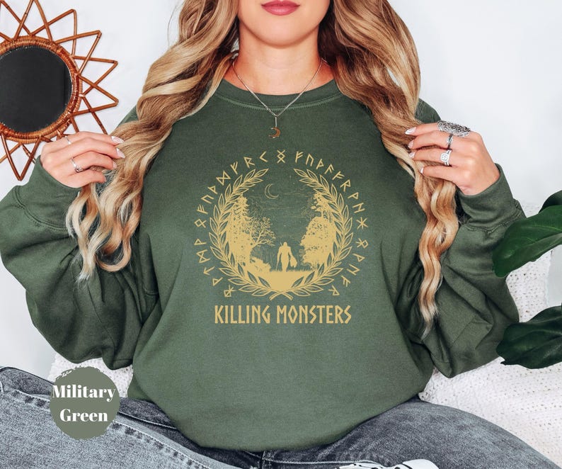 May include: Military green sweatshirt with a gold graphic featuring a forest scene, a crescent moon, and the text "KILLING MONSTERS". The design is encircled by a wreath and runic symbols. The sweatshirt is a solid color.