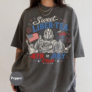May include: Dark gray t-shirt with a patriotic graphic. The design features a soldier giving a thumbs up, the Statue of Liberty, an American flag, and the text "Sweet Liber-Tea est 1776 4th of July Club."