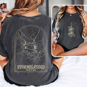 May include: Pepper-coloured t-shirt with a graphic on the back featuring a storm scene with lightning and the text "STORMBLESSED THE SKY IS MINE." The front of the shirt has a spear graphic with the text "BEFORE DESTINATION."