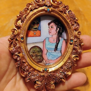 May include: Ornate gold-colored oval frame with a miniature painting inside. The painting depicts a woman in a blue dress and a creature with an orange face. The frame has blue accents and is held in a hand.