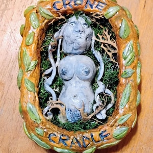 Puede incluir: Escultura hecha a mano de una figura femenina dentro de un marco. La figura es azul claro, con un rostro y torso detallados. El marco es marrón con hojas verdes y las palabras "CRONE" y "CRADLE" en azul.