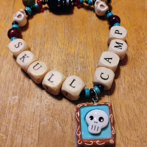 May include: A handmade bracelet with a skull theme. The bracelet features wooden cube beads spelling "SKULL CAMP", skull-shaped beads, and a pendant with a white skull on a turquoise background. The beads are strung with black and turquoise accents.