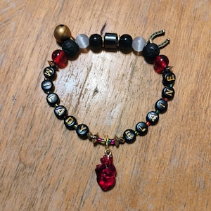 May include: A beaded bracelet featuring black letter beads spelling MILADY ELAINE, interspersed with black, red, and translucent white beads. It includes a gold-toned bell, a horseshoe charm, and a central red anatomical heart-shaped pendant.