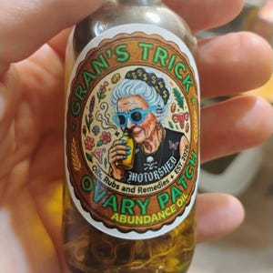 Può includere: Una bottiglia di vetro trasparente riempita con olio color ambra e pezzi botanici. L'etichetta presenta l'illustrazione di una persona anziana con capelli bianchi, occhiali blu e maglietta nera, con il testo Gran's Trick Ovary Patch Abundance Oil.