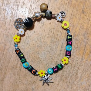 May include: A beaded bracelet with the words "ATOMIC GOAT SHE" spelled out in colorful letter beads. The bracelet features a black 8-ball bead, flower beads, and a sun charm. Other beads are silver and brown.