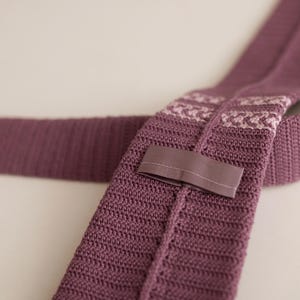 May include: A knitted tie in a muted purple hue. The tie features a textured knit pattern and a contrasting patterned section near the knot. A fabric loop is sewn on the back. The tie is displayed on a light background.