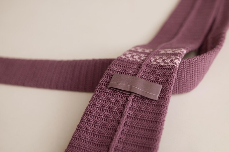 May include: A knitted tie in a muted purple hue. The tie features a textured knit pattern and a contrasting patterned section near the knot. A fabric loop is sewn on the back. The tie is displayed on a light background.